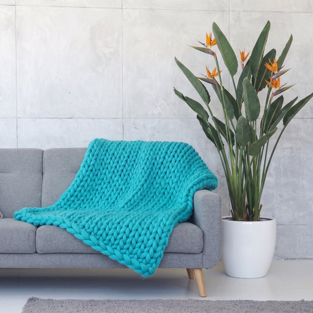Plaid en grosse maille tricoté main en laine mérinos XXL coloris Turquoise lagon, posé sur un canapé gris à côté d’un oiseau de paradis