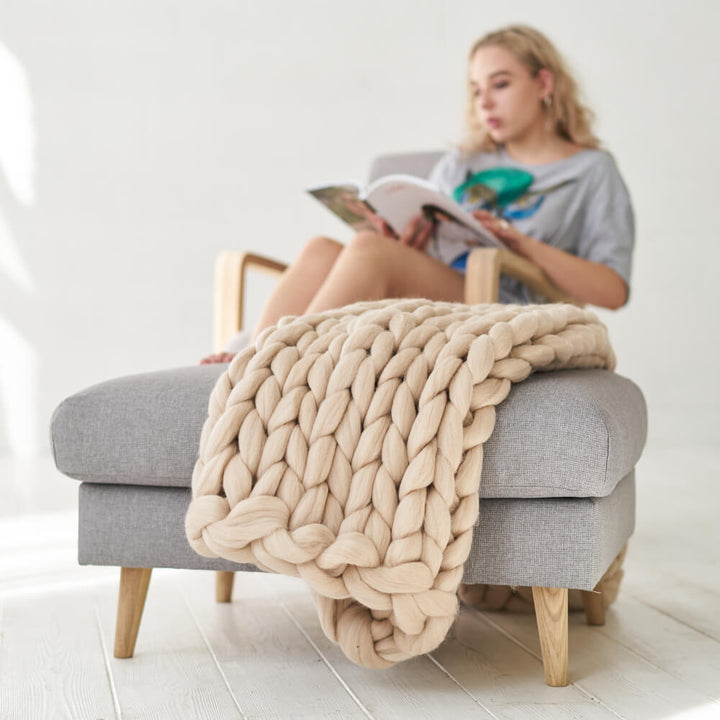 beige chunky knit lap blanket draped over grey ottoman and girls is sitting on the chair and reading a magazine