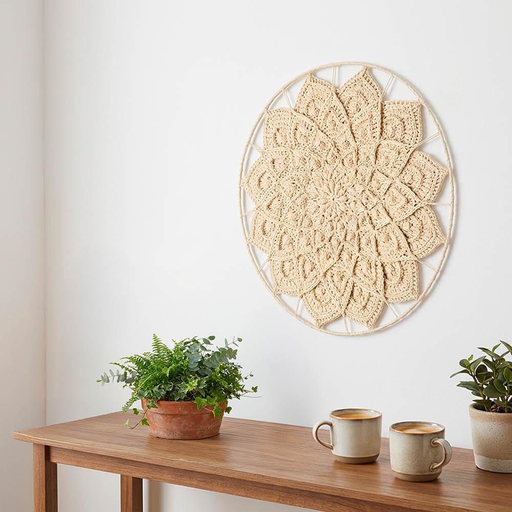 Decorative wall art with a floral pattern on a white wall above a wooden table with plants and mugs.
