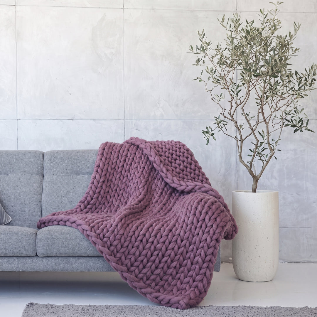 Plaid en grosse maille tricoté main en laine mérinos XXL coloris Prune, posé sur un canapé gris à côté d’un olivier