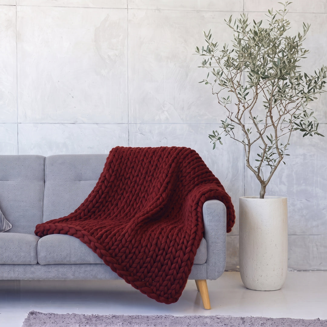 Plaid en grosse maille tricoté main en laine mérinos XXL coloris Baiser bordeaux, posé sur un canapé gris à côté d’un olivier