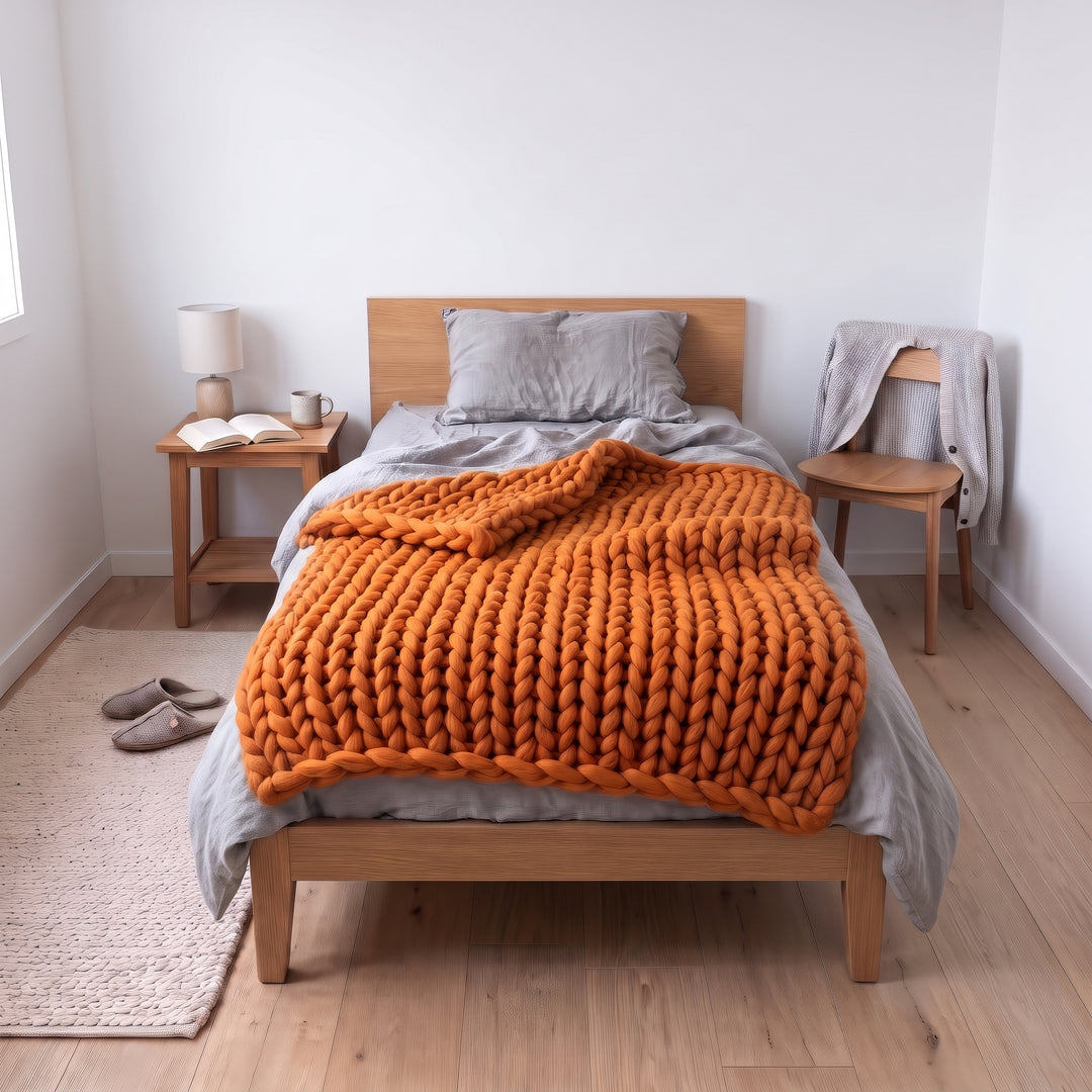 Grob gestrickte Wolldecke aus Merinowolle in der Farbe Butternusskürbis, locker auf einem Einzelbett mit grauer Leinenbettwäsche in einem hellen Schlafzimmer