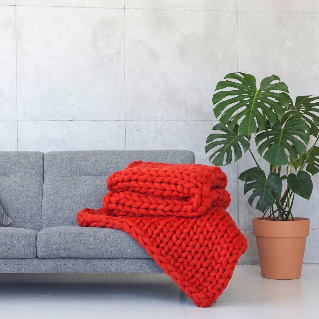 Plaid en grosse maille tricoté main en laine mérinos XXL coloris Rouge bonbon, plié sur un canapé gris à côté d’un monstera