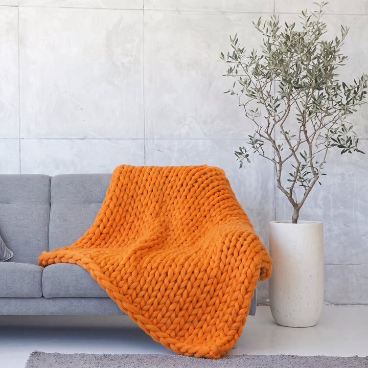 Plaid en grosse maille tricoté main en laine mérinos XXL coloris Roux renard, posé sur un canapé gris à côté d’un olivier