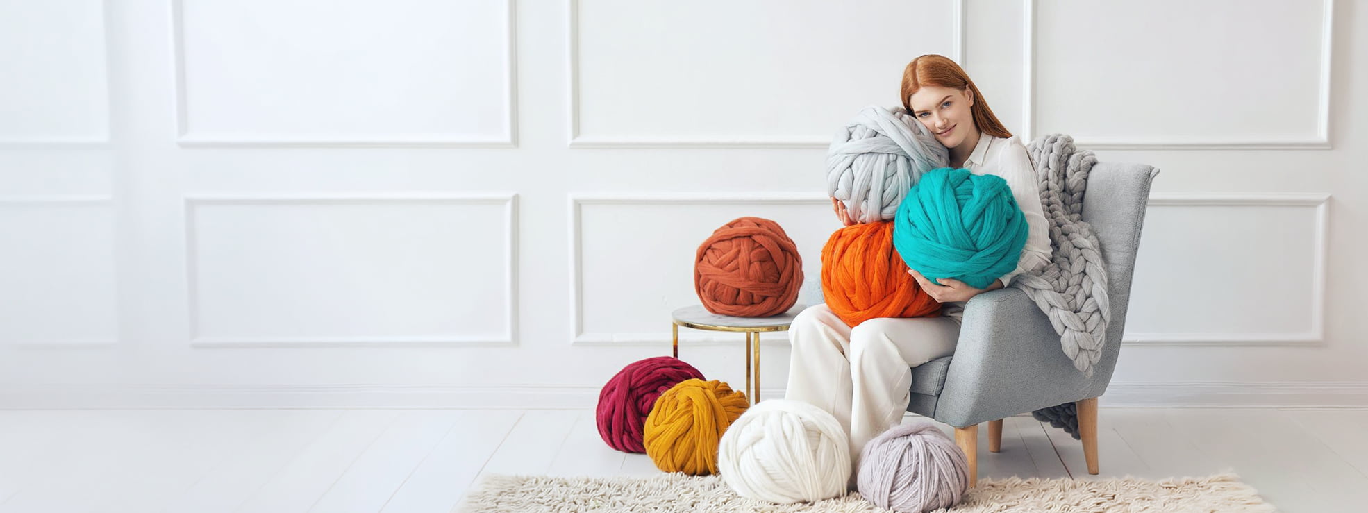 Woman hugging chunky merino wool yarn balls in grey and teal, surrounded by orange, mustard, burgundy and white chunky yarns — handmade by Cheery Little Sheep