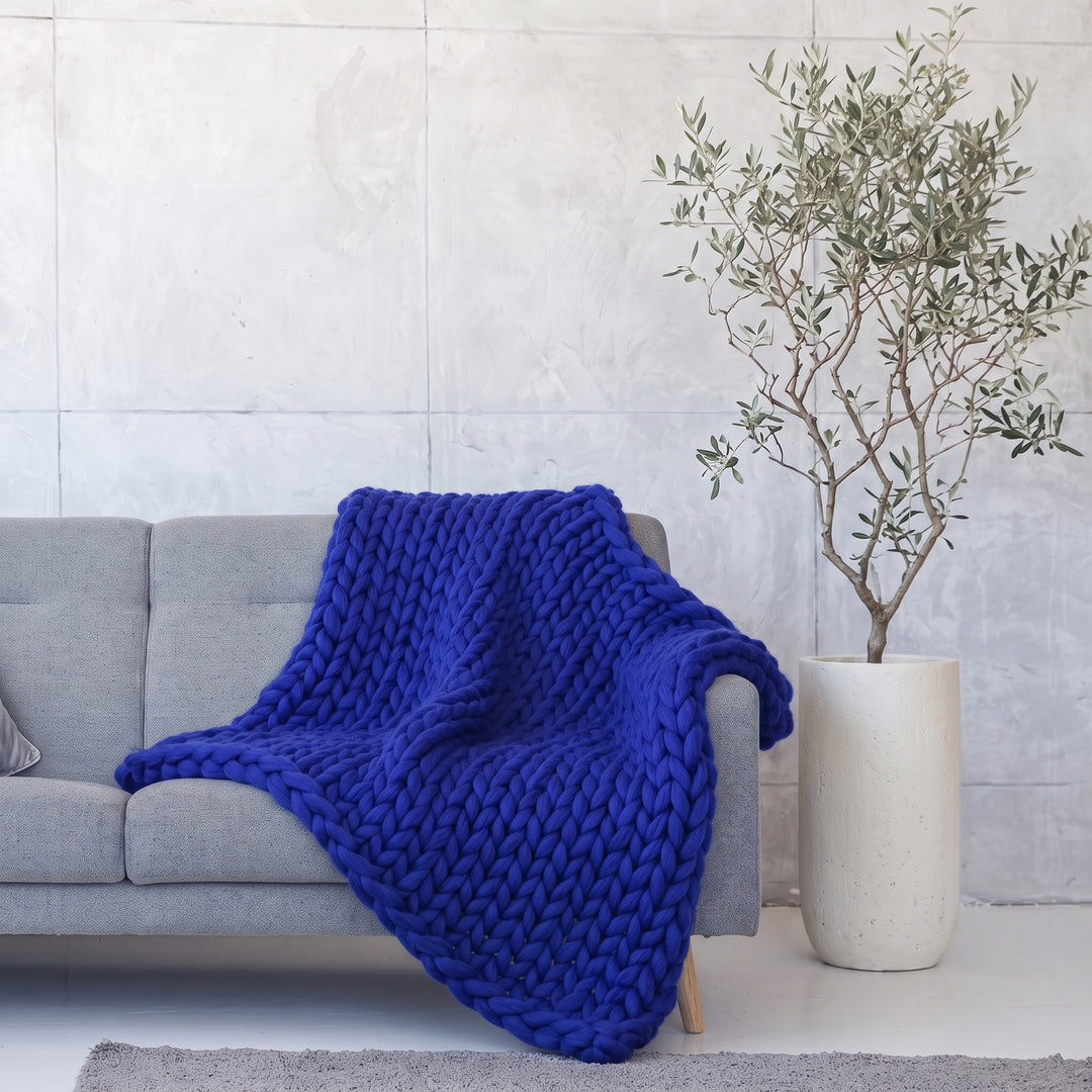 Plaid en grosse maille tricoté main en laine mérinos XXL coloris Bleu cobalt, posé sur un canapé gris à côté d’un olivier