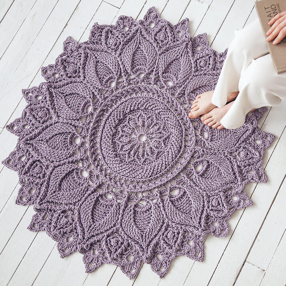 Crocheted purple round rug on a white wooden floor with a person sitting on it.