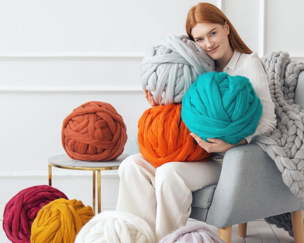 girl sitting on chair and halding large balls of various colour merino wool chunky yarn