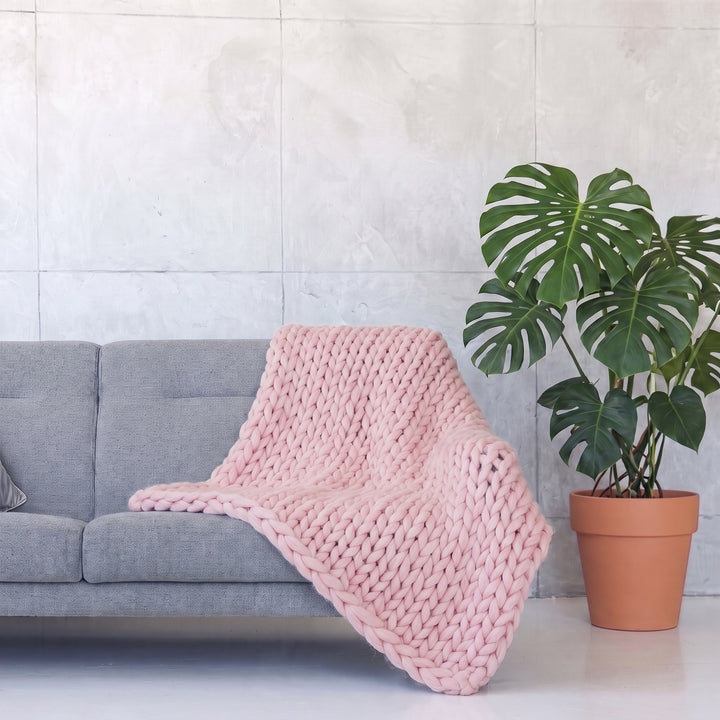 Plaid en grosse maille tricoté main en laine mérinos XXL coloris Rose pâle, posé sur un canapé gris à côté d’un monstera