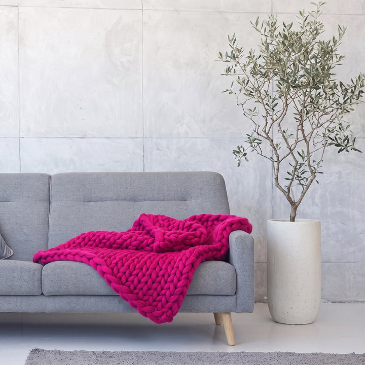 Plaid en grosse maille tricoté main en laine mérinos XXL coloris Fuchsia vif, posé sur un canapé gris à côté d’un olivier