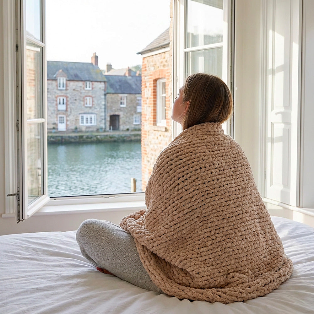 girl wrapped in a chenille blanket on the bed looking through the open window