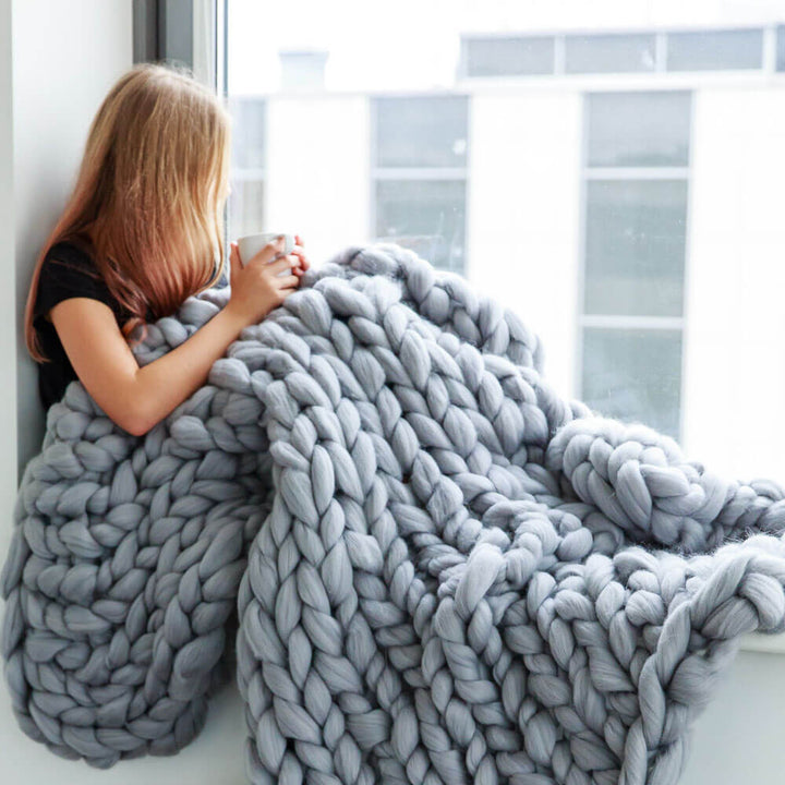 girl wrapped in grey chunky knit blanket sitting at the window