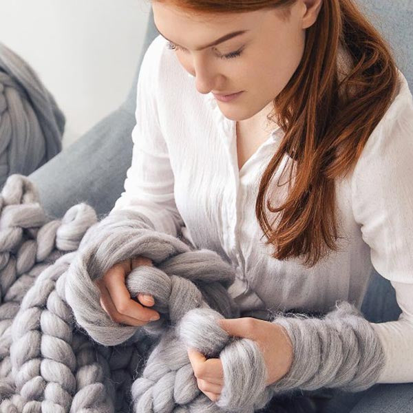 Woman holding a large gray knitted blanket in a cozy indoor setting.