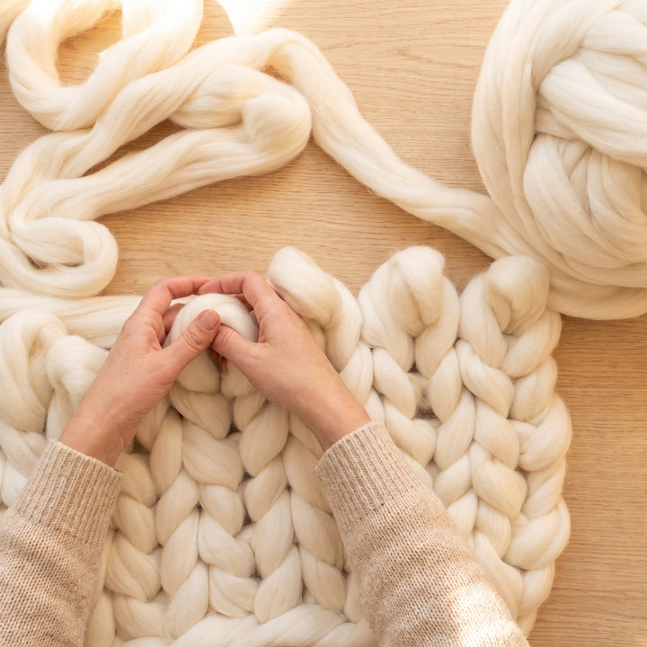 Hände in einem gemütlichen haferfarbenen Pullover stricken eine Grobstrickdecke aus cremeweißer dicker Merinowolle auf einem hellen Eichentisch, Ansicht von oben