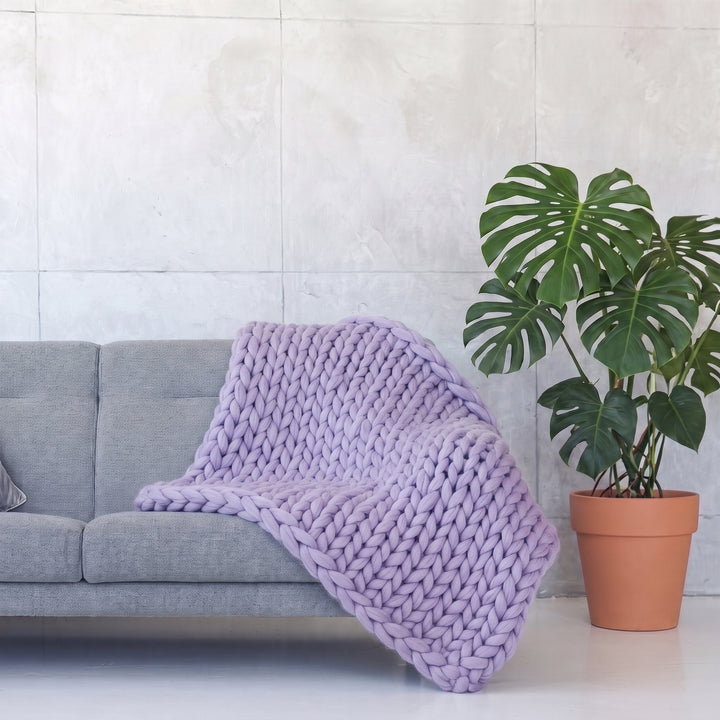 Plaid en grosse maille tricoté main en laine mérinos XXL coloris Lavande, posé sur un canapé gris à côté d’un monstera