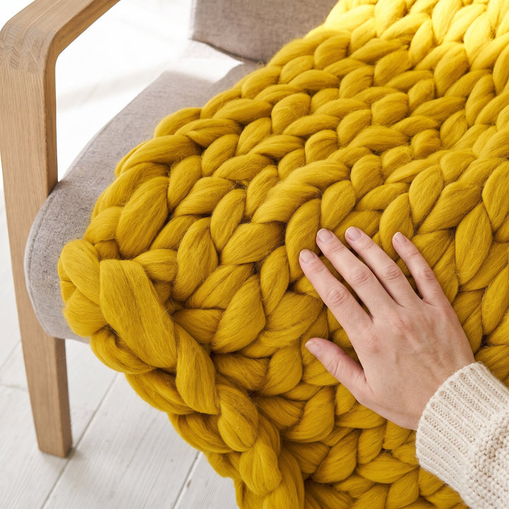 Nahaufnahme einer senfgelben grob gestrickten Decke aus Merinowolle mit einer Hand, die die Größe der Maschen zeigt