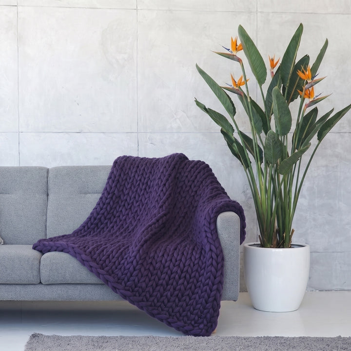 Plaid en grosse maille tricoté main en laine mérinos XXL coloris Indigo mystérieux, posé sur un canapé gris à côté d’un oiseau de paradis