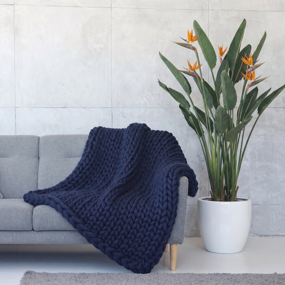 Plaid en grosse maille tricoté main en laine mérinos XXL coloris Bleu Oxford, posé sur un canapé gris à côté d’un oiseau de paradis