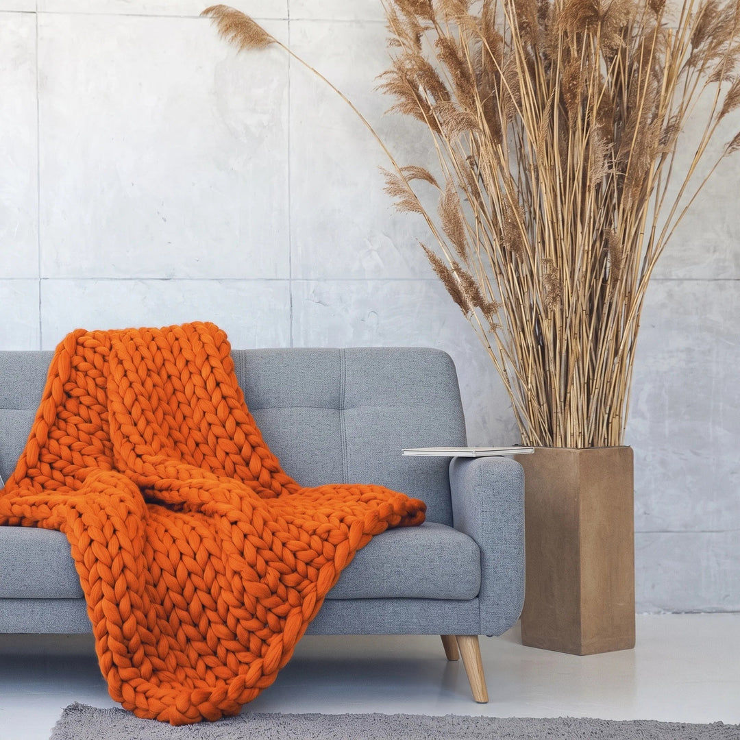 Plaid en grosse maille tricoté main en laine mérinos XXL coloris Orange feu, posé sur un canapé gris à côté d’herbes de la pampa séchées