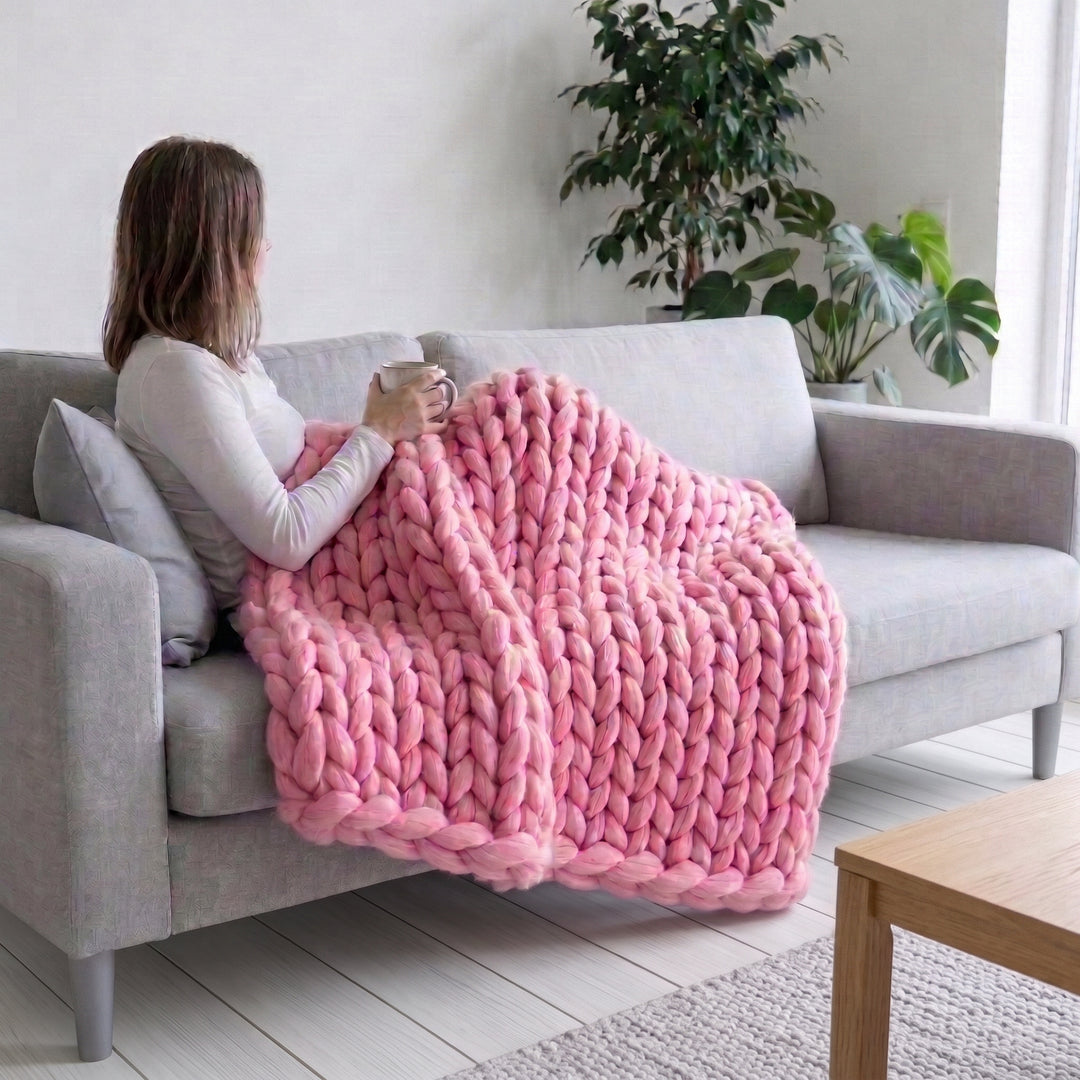 Frau auf einem hellgrauen Sofa mit einer Tasse, die Beine mit einer rosa grob gestrickten Wolldecke aus Merinowolle zugedeckt
