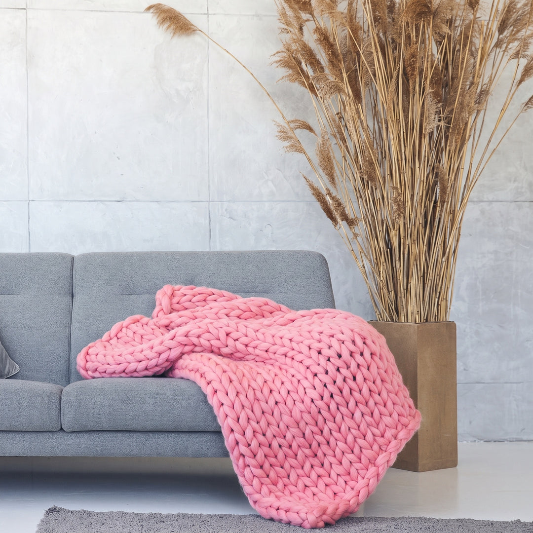 Plaid en grosse maille tricoté main en laine mérinos XXL coloris Rose limonade, posé sur un canapé gris à côté d’herbes de la pampa séchées