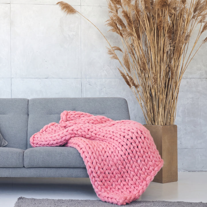 Plaid en grosse maille tricoté main en laine mérinos XXL coloris Rose limonade, posé sur un canapé gris à côté d’herbes de la pampa séchées
