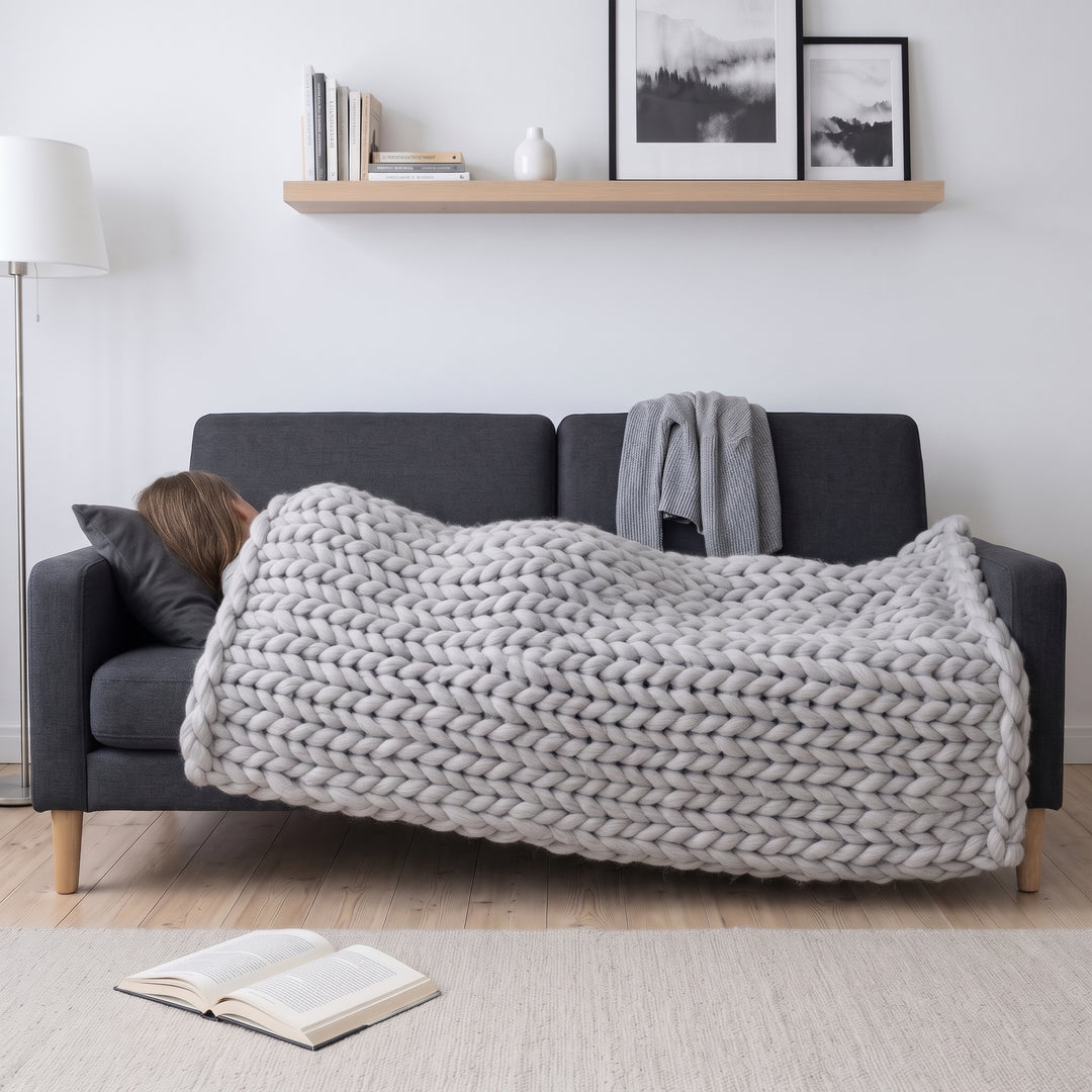 Frau schläft auf einem dunkelgrauen Sofa, von den Schultern bis zu den Füßen zugedeckt mit einer hellgrauen grob gestrickten Wolldecke aus Merinowolle in der Farbe Regenzeit