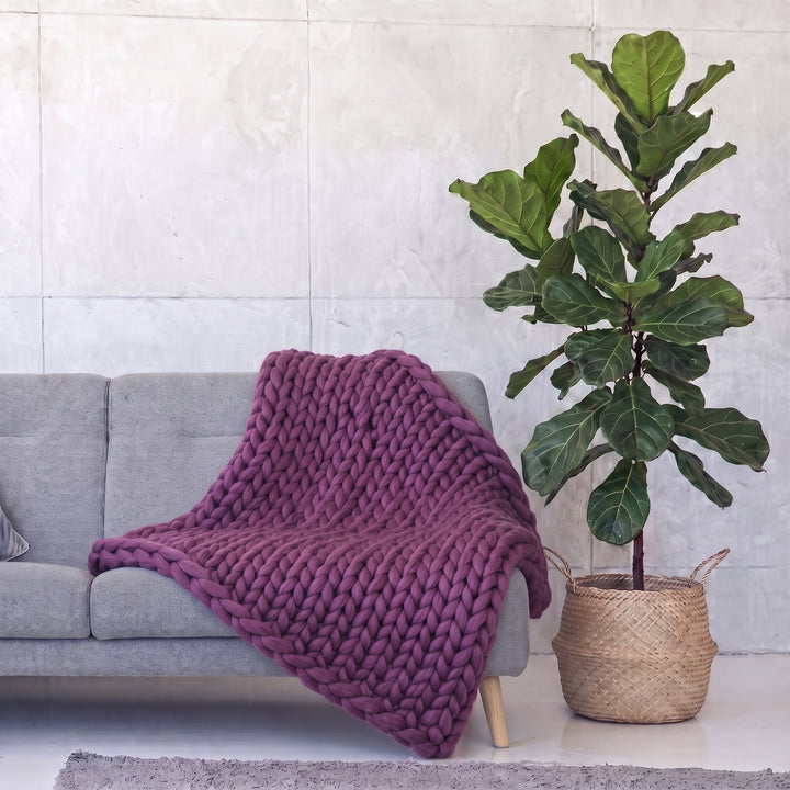 Plaid en grosse maille tricoté main en laine mérinos XXL coloris Raisin doux, posé sur un canapé gris à côté d’un figuier lyre