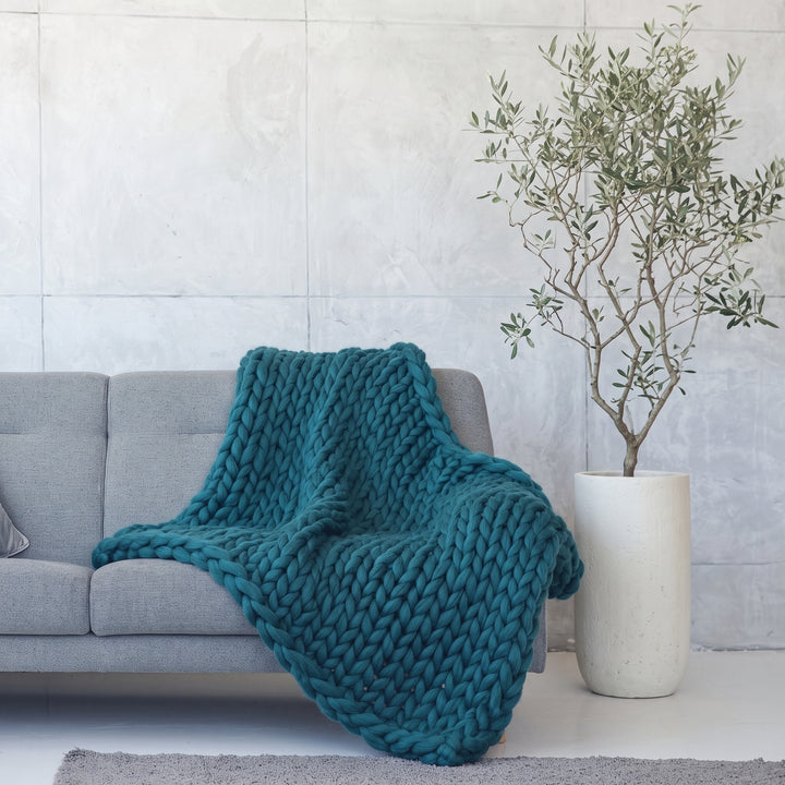 Plaid en grosse maille en laine mérinos tricoté à la main, coloris Bleu canard, sur un canapé gris à côté d’un olivier