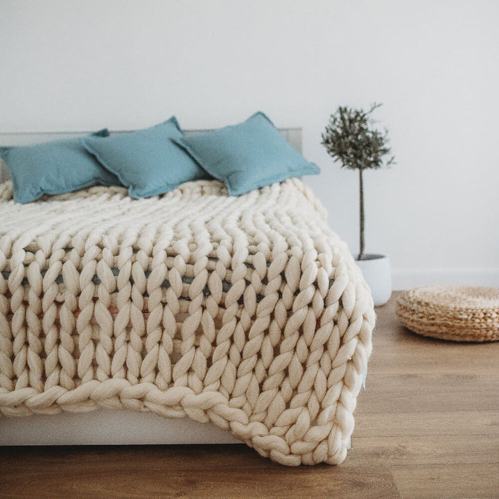 a white chunky wool blanket on the bed with blue pillows and plant at the bed