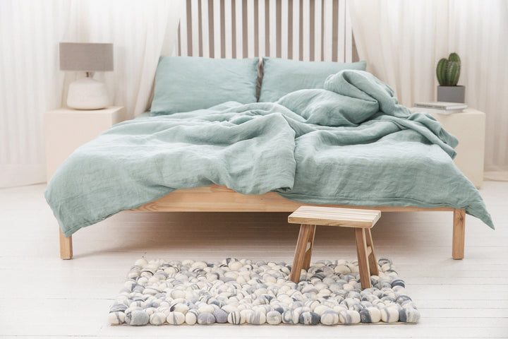 a bedroom with bed and green bedding, felted wool pebbles rug at the feet of the bed and a littles stool on it